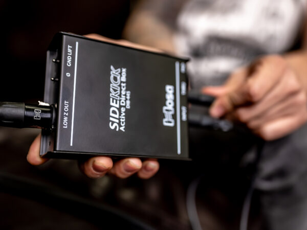 A musician holds the Hosa DIB-445 Sidekick Active DI Box in their hand, connecting an instrument cable to the input.